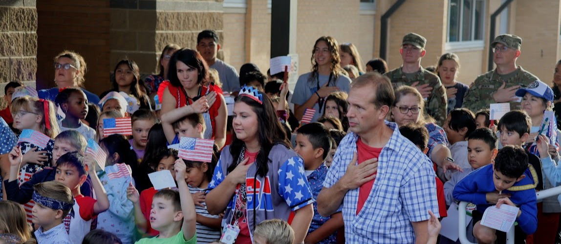 A crowd of people, including children, adults, and uniformed individuals, gather outdoors. Some hold American flags.