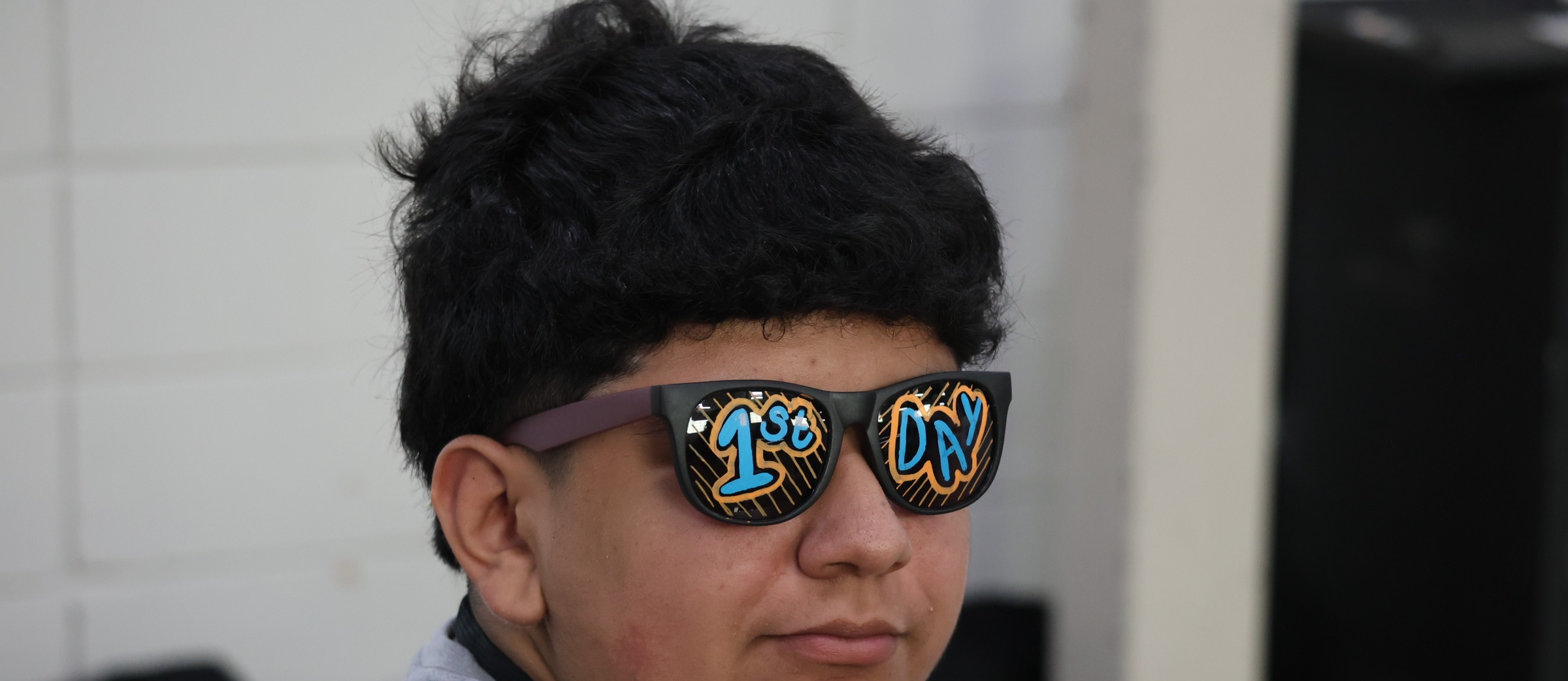 Person with dark hair, wearing a gray shirt and sunglasses with "1st" and "UN" printed on them.