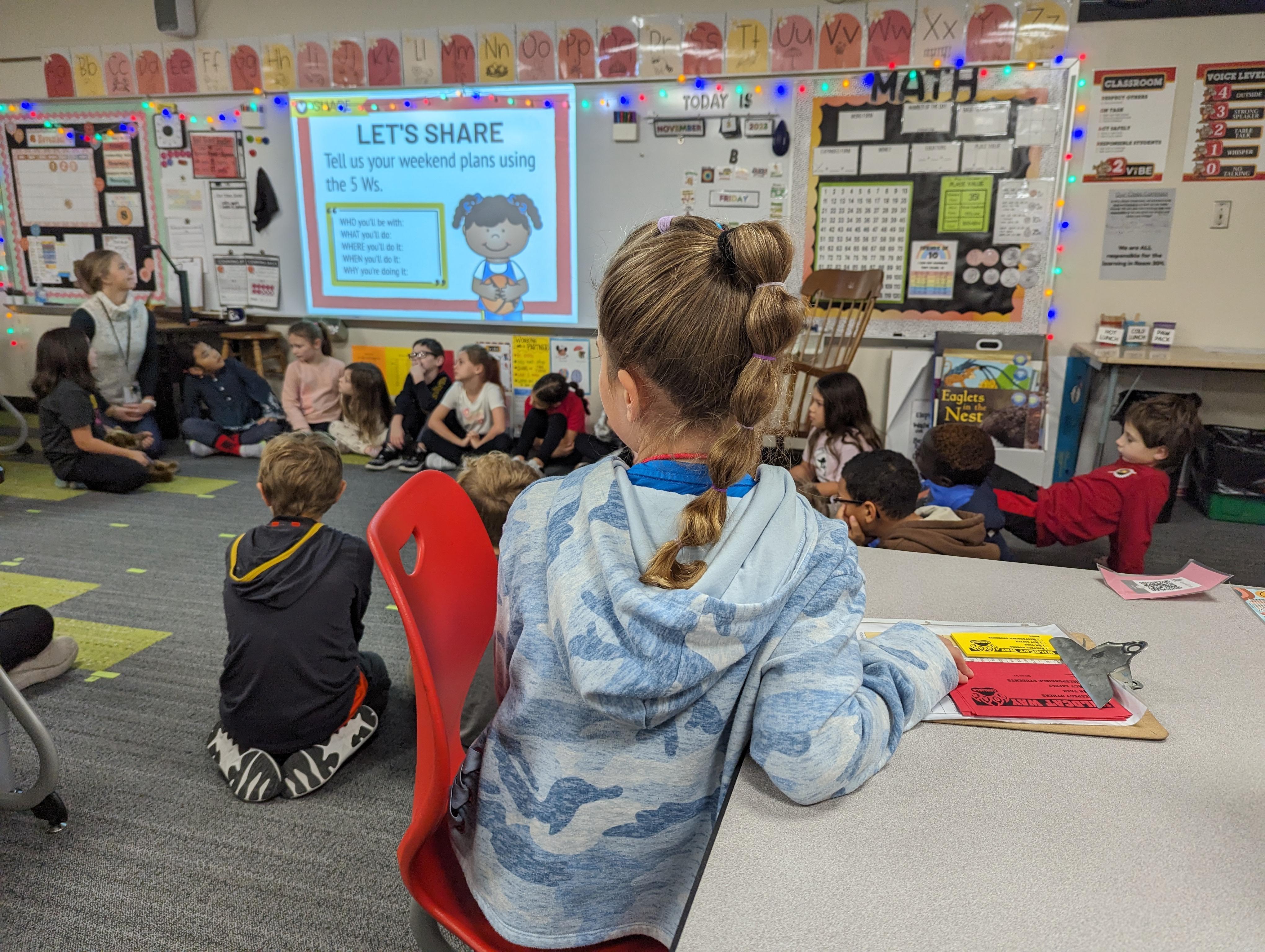 Child sitting at a desk in front of a teacher, attentively listening to a presentation with other students around them.