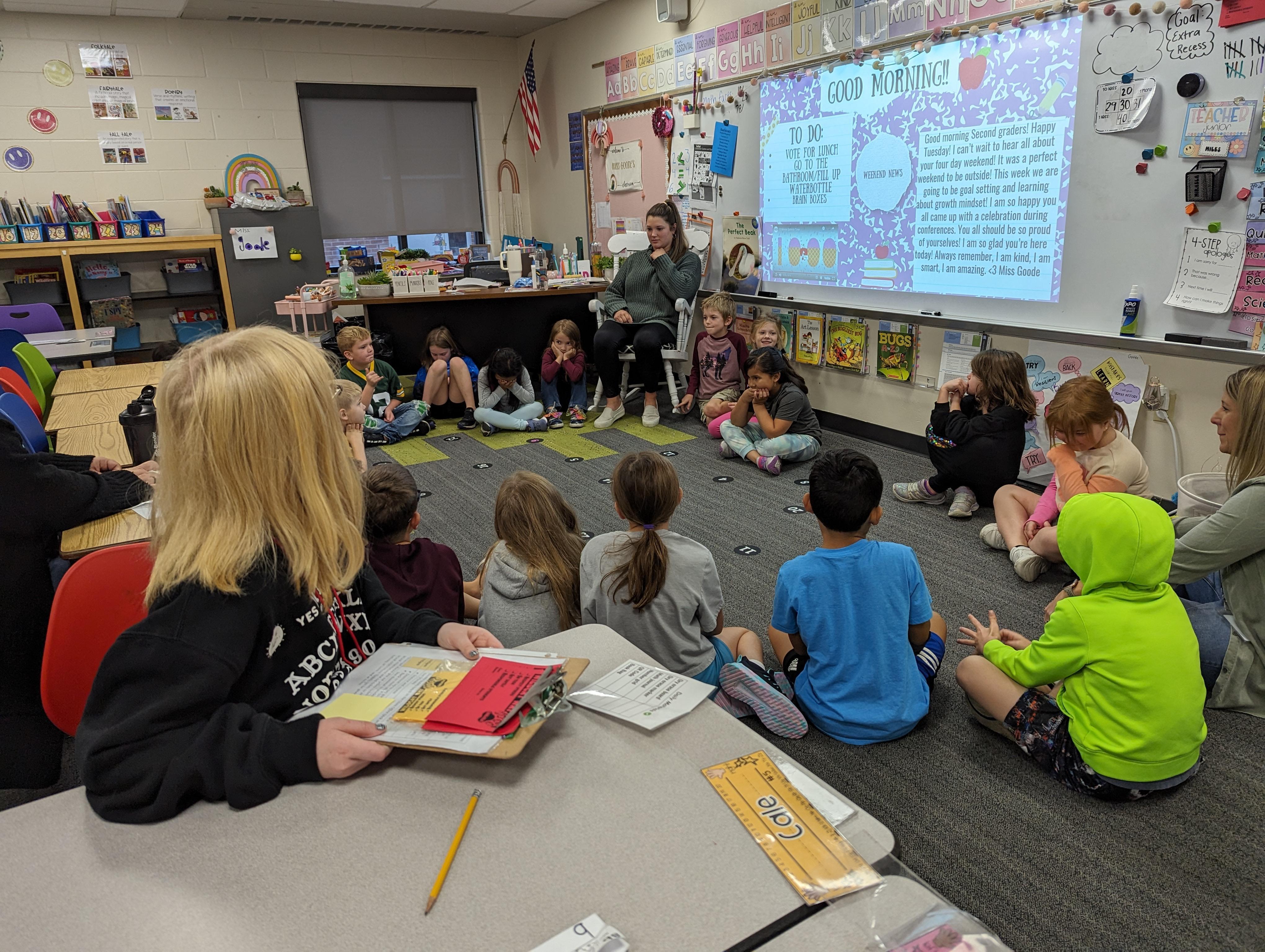 Children in a classroom, gathered around a teacher presenting on a whiteboard.