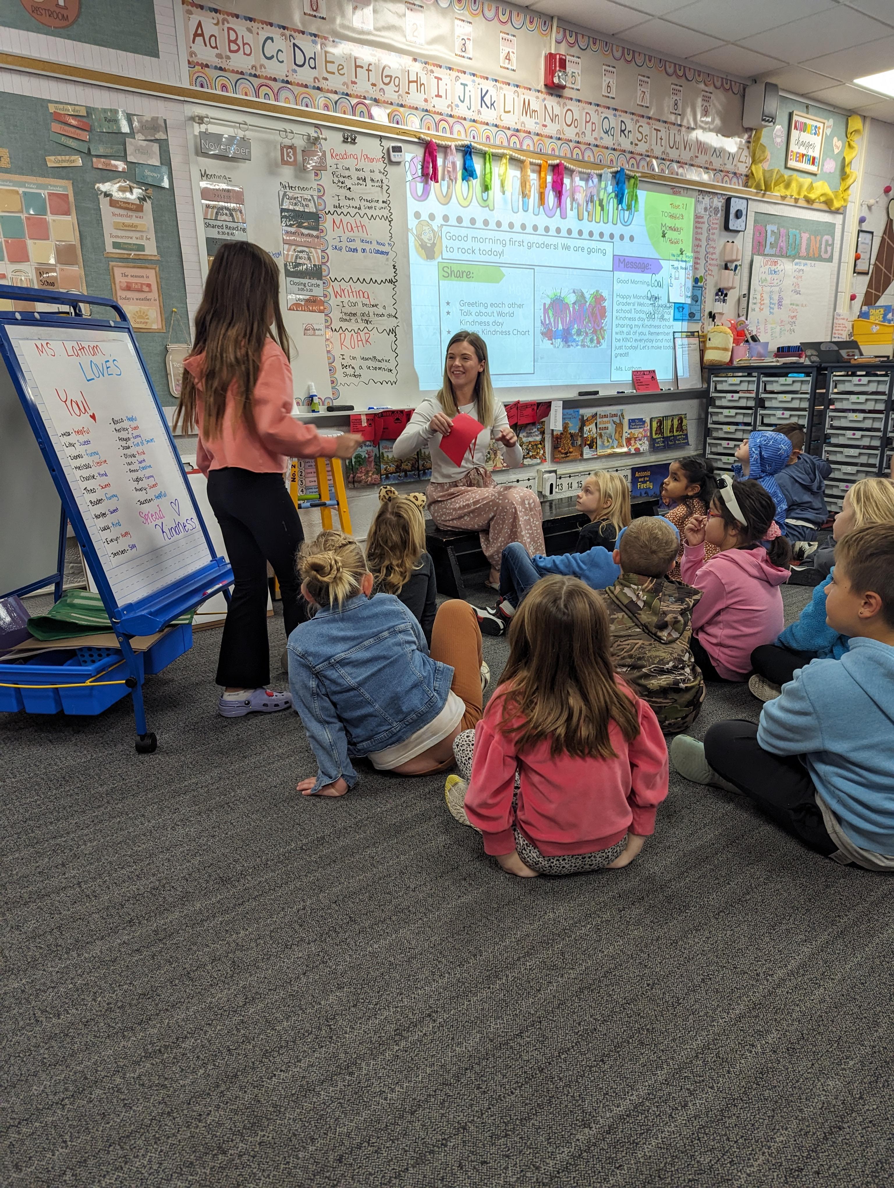 A teacher and a student in a classroom setting, with the teacher presenting information from a whiteboard to children seated on the floor.