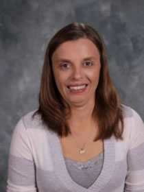 A woman wearing a cozy gray sweater over a crisp white shirt, smiling warmly at the camera.