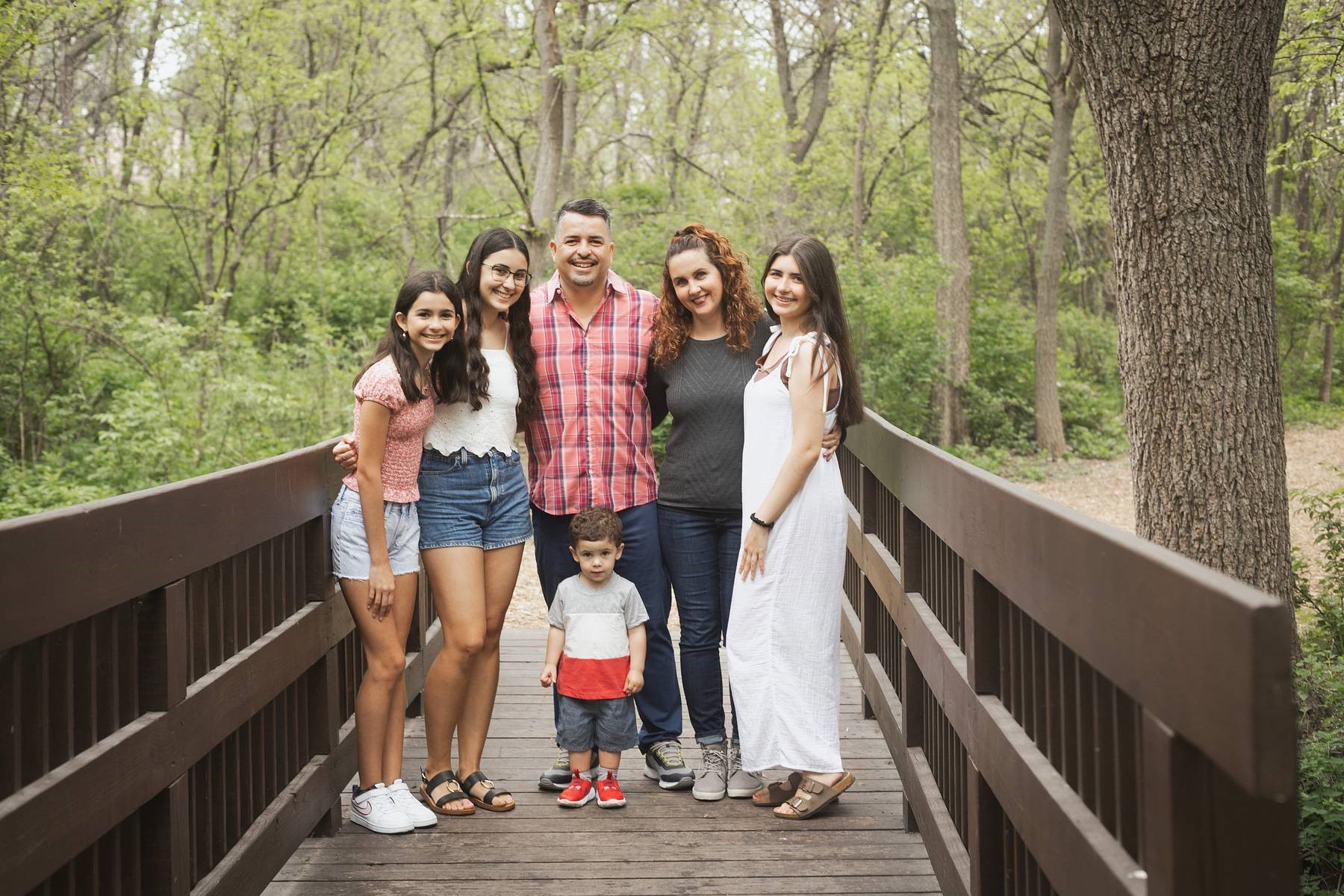 A family smiling on a bridge in the woods, surrounded by lush green trees and a serene river flowing underneath.