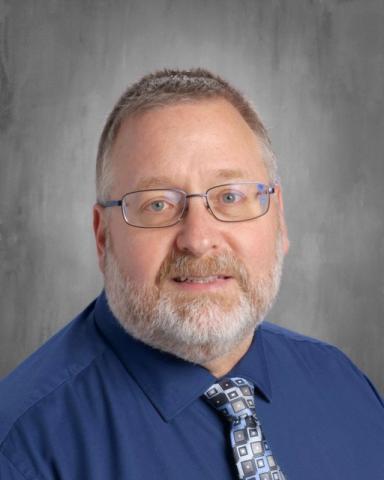 A man in a blue shirt and tie stands against a gray background. He wears glasses, has a beard, and smiles.
