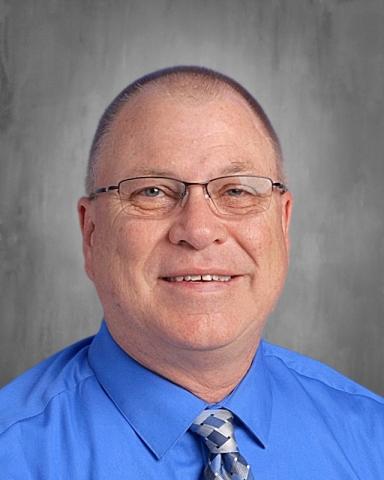 A man wearing a blue shirt and tie with a smile, set against a plain gray background.