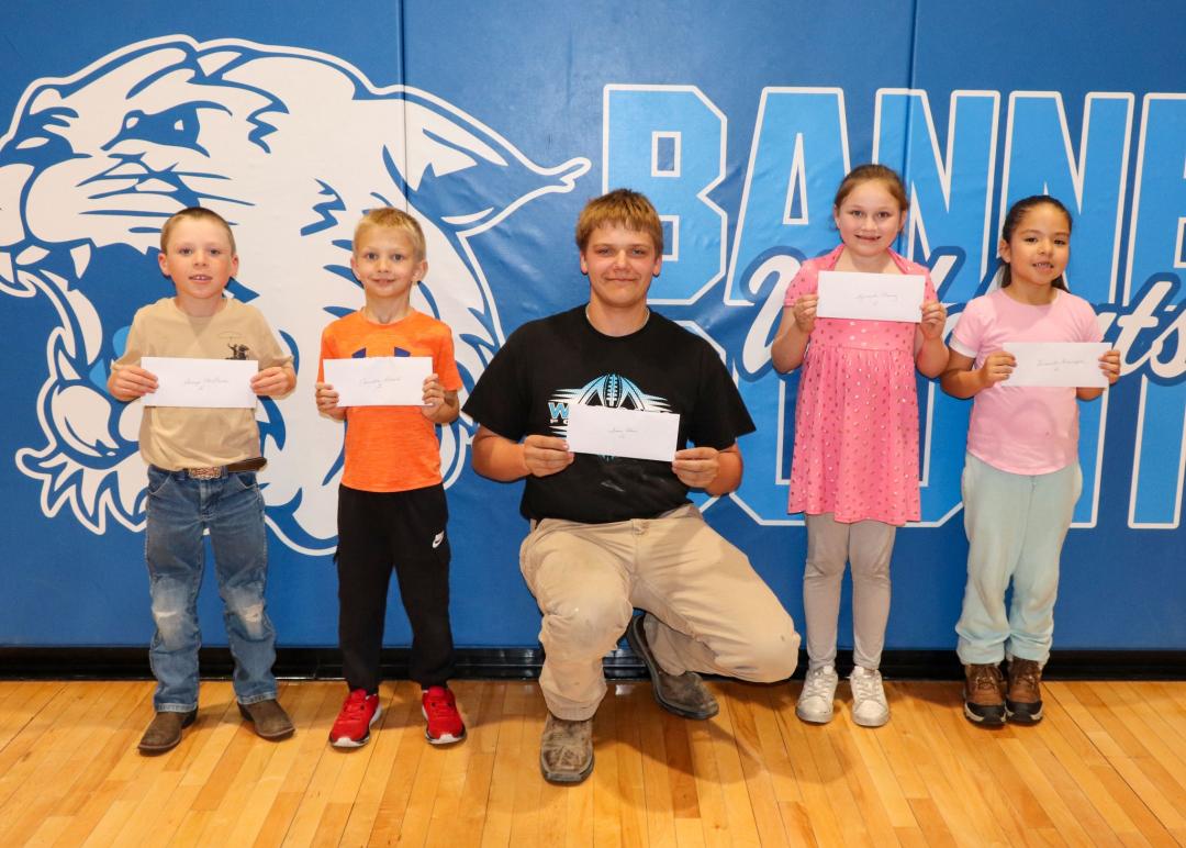 Five children holding papers pose with a kneeling adult in front of a blue wall with "BANNER" text.