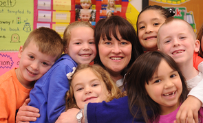 A woman embraces a group of children in a classroom setting.