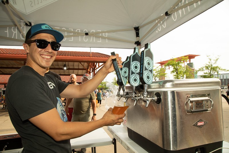 A man in sunglasses pours beer into a machine in West Allis, WI, capturing a fun moment of brewing excitement.