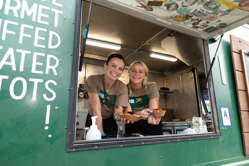 Two women serve delicious food from vibrant green food trucks in West Allis, WI, creating a lively culinary scene.