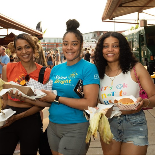 Three women joyfully holding delicious food at an outdoor market in West Allis, WI, surrounded by vibrant stalls and shoppers.