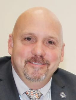 A close-up portrait of a bald man with a goatee, wearing a suit and tie, smiling at the camera.