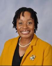 A woman with short, curly hair wearing a yellow blazer and a pearl necklace smiles at the camera against a light gray background.