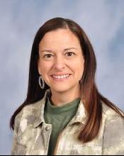 A woman with long brown hair, wearing a green shirt and a patterned jacket, smiles at the camera against a soft blue background.