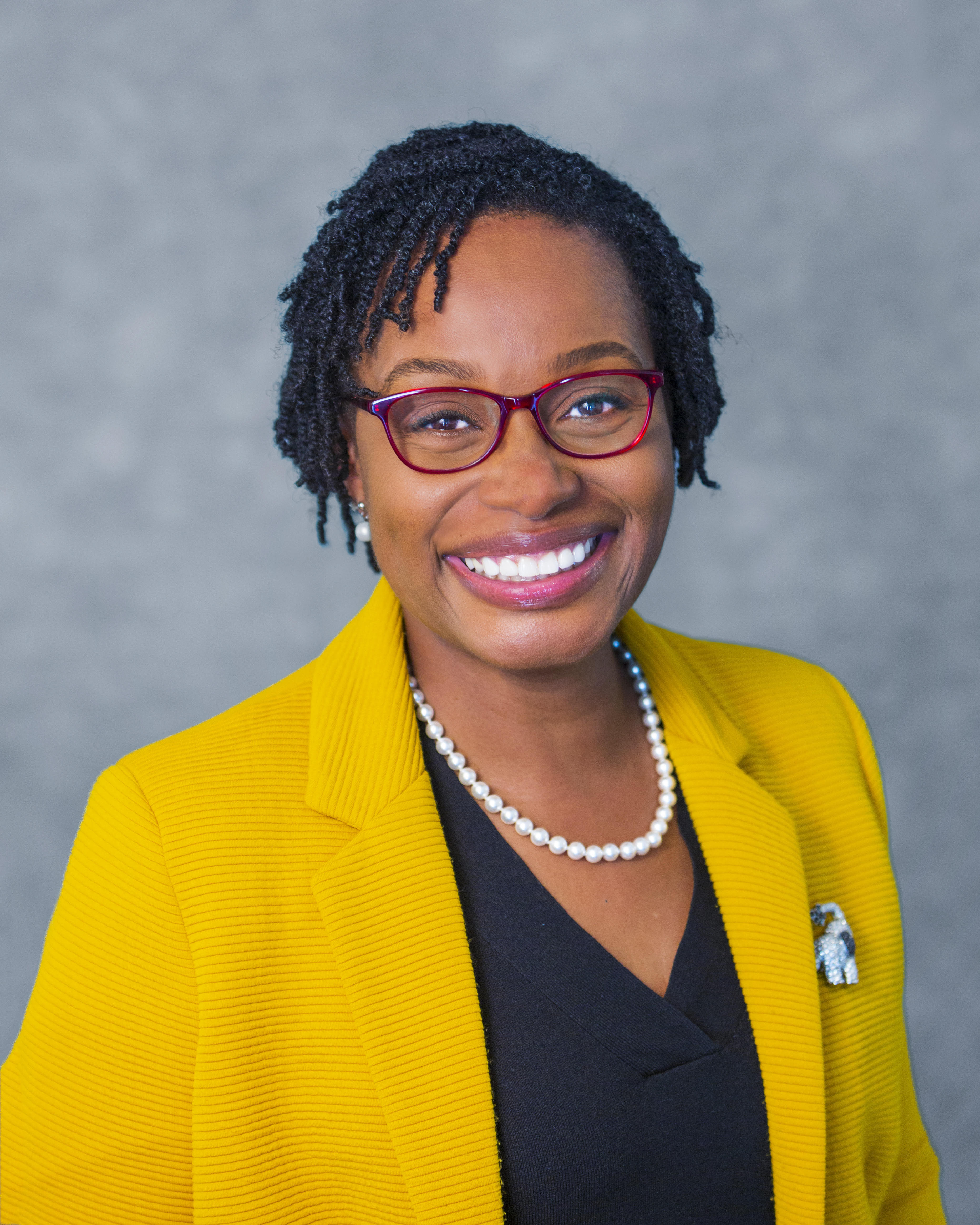 A smiling woman with short, curly hair wearing glasses, a yellow blazer, and a pearl necklace, set against a gray background.