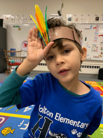 A young boy in a classroom wearing a colorful feather headband, engaged in a creative learning activity.