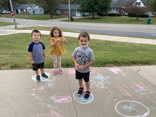 Three children standing in front of colorful chalk drawings on the sidewalk.