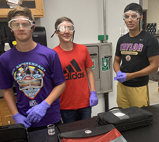 Three young men in lab coats and goggles standing next to a table in a laboratory setting.