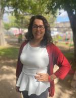 A woman wearing glasses and a white shirt stands confidently in front of a lush green tree, showcasing a serene outdoor setting.