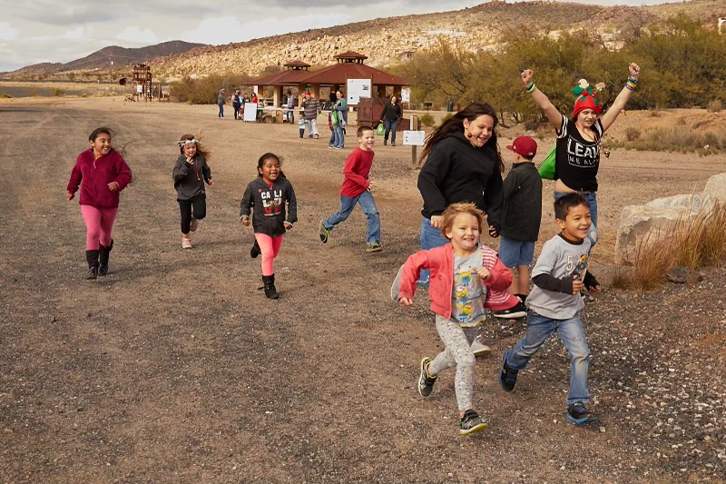 A group of joyful children running together across a vast, sandy desert under a bright blue sky.
