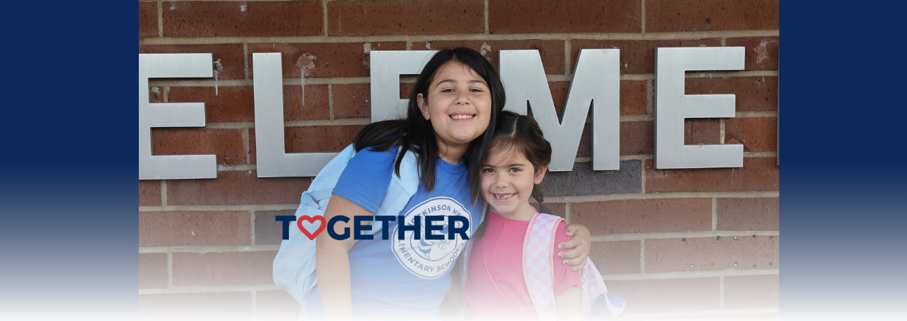 Two HHES students smile and hug one another in front of the school building.