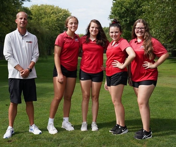 Four girls wearing red shirts and black shorts pose together on a golf course, showcasing a vibrant and sporty atmosphere.