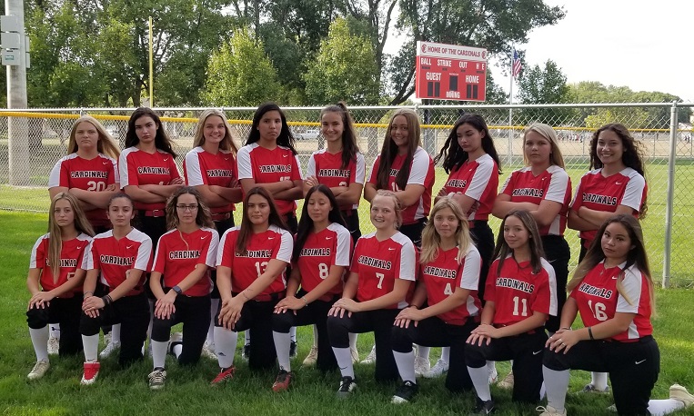 A group of softball players smiling and posing for a team photo on the field.