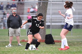 wo girls playing soccer on a field with a crowd watching.
