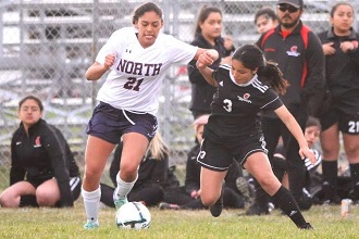 Two young female soccer players sprinting towards the ball on the field.