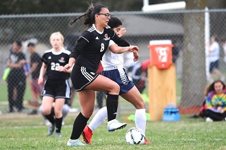 A girl in black soccer uniform kicking the ball on the field.