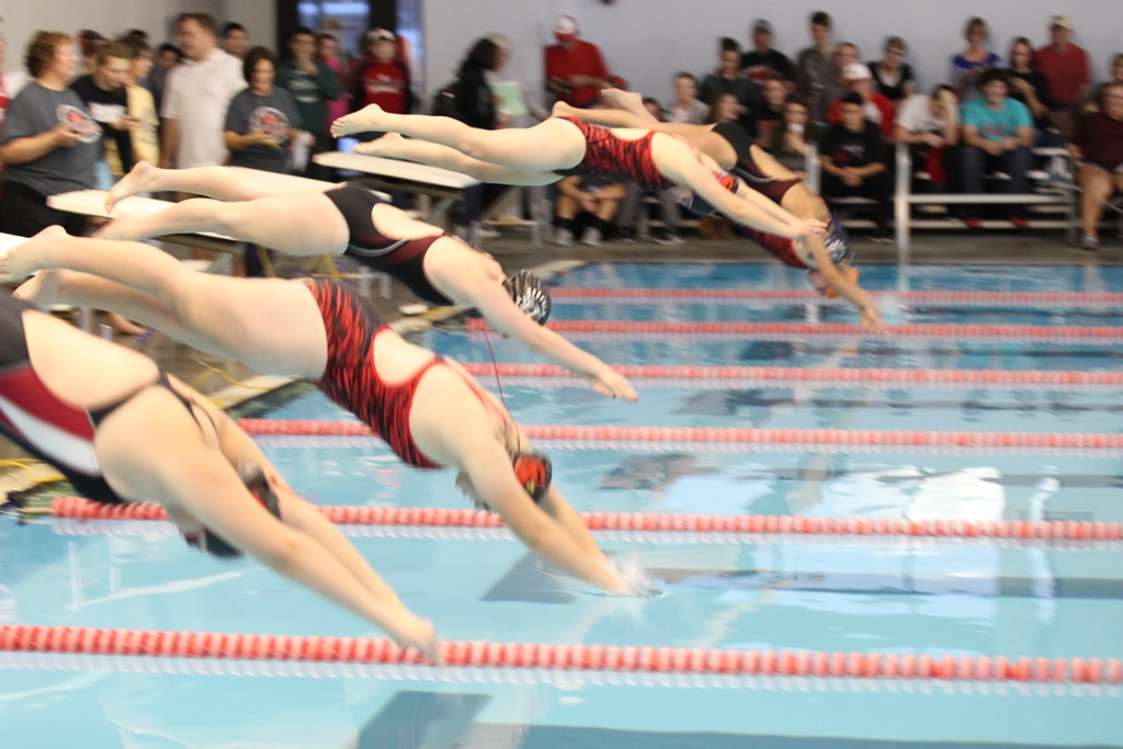 Swimmers diving into pool during competition, creating ripples in water.