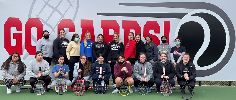 A group of young women smiling and posing with tennis rackets outdoors.