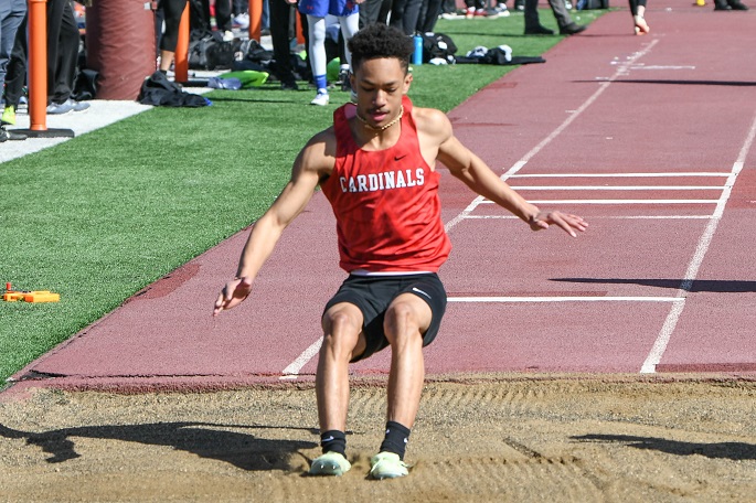 A young man in a red shirt leaps into the air during a long jump event, showcasing athleticism and determination.
