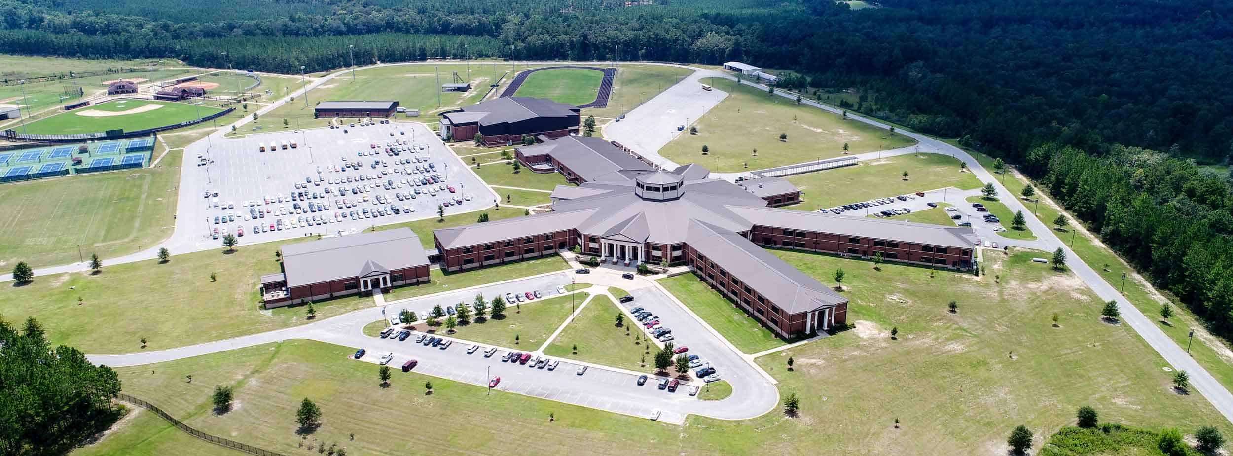 Aerial view of a large high school campus with a central circular building, multiple parking lots, sports fields, tennis courts, and surrounding wooded areas.