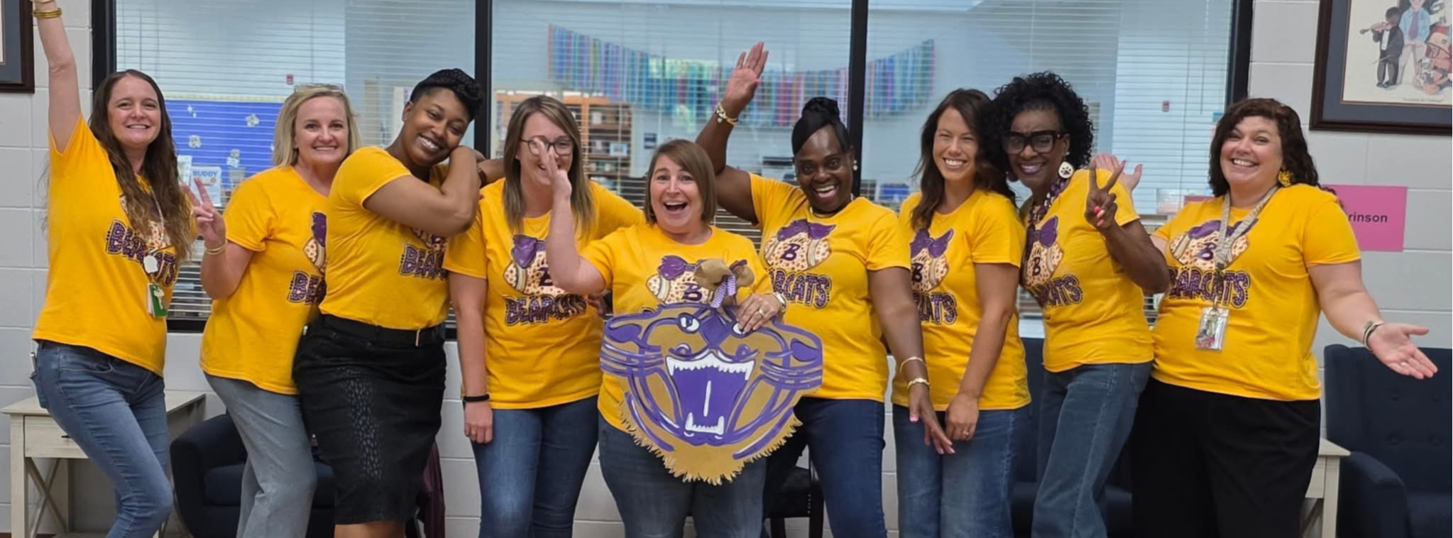 A group of nine smiling women wearing matching gold “Bearcats” t-shirts pose together indoors, holding a large cutout of a purple bearcat mascot.