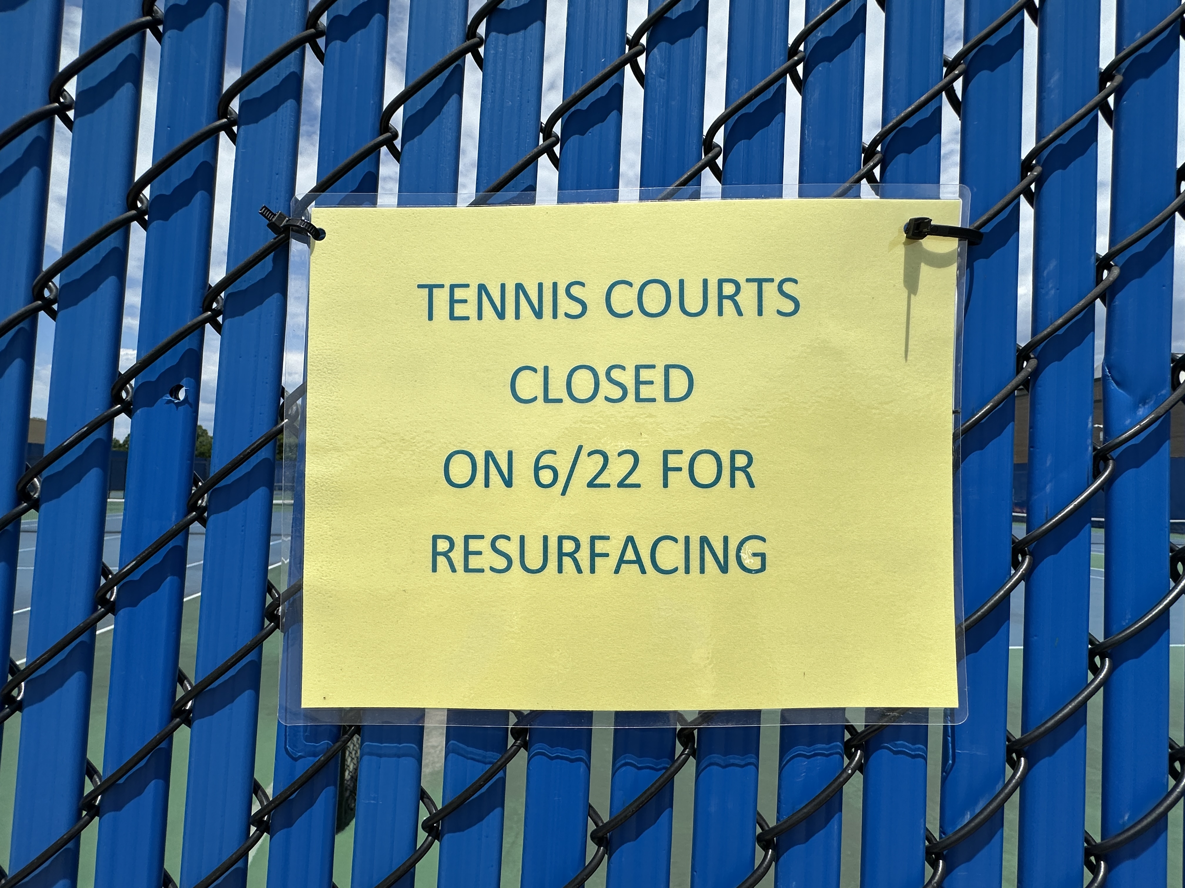 A sign on a fence that reads, "Tennis Courts Close on 6/22 for Resurfacing."