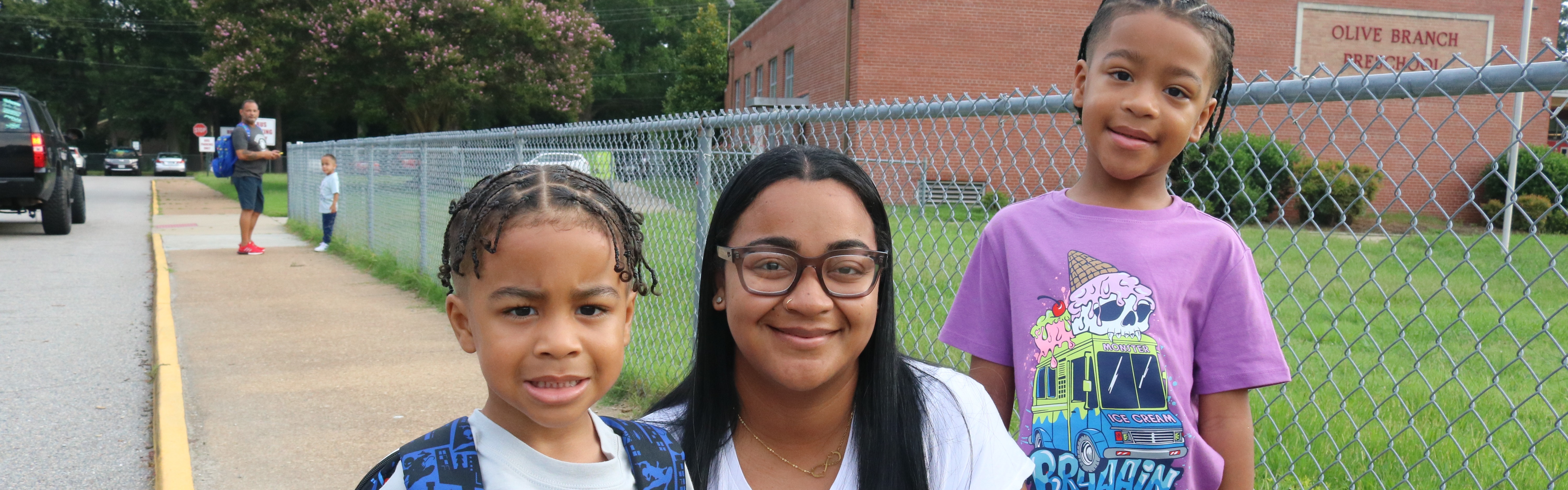 A woman and two children stand together in front of a fence and building. One child wears a backpack.