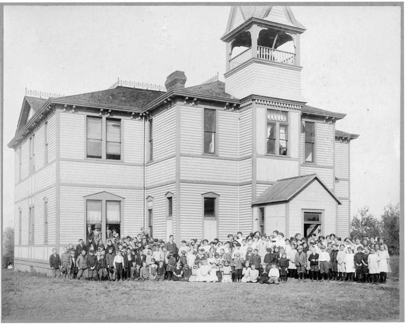 An old black and white photo depicting a group of people gathered in front of a school building, showcasing historical attire.