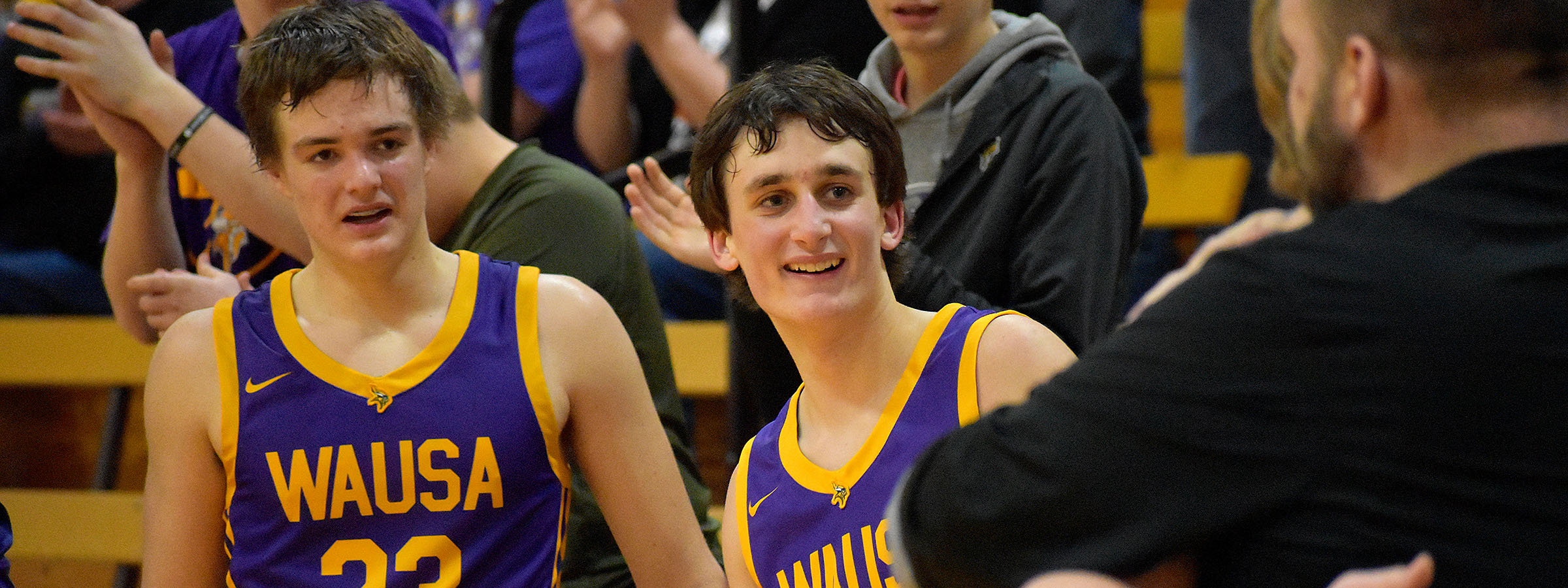 Two boys in purple and gold basketball uniforms smiling on the court.