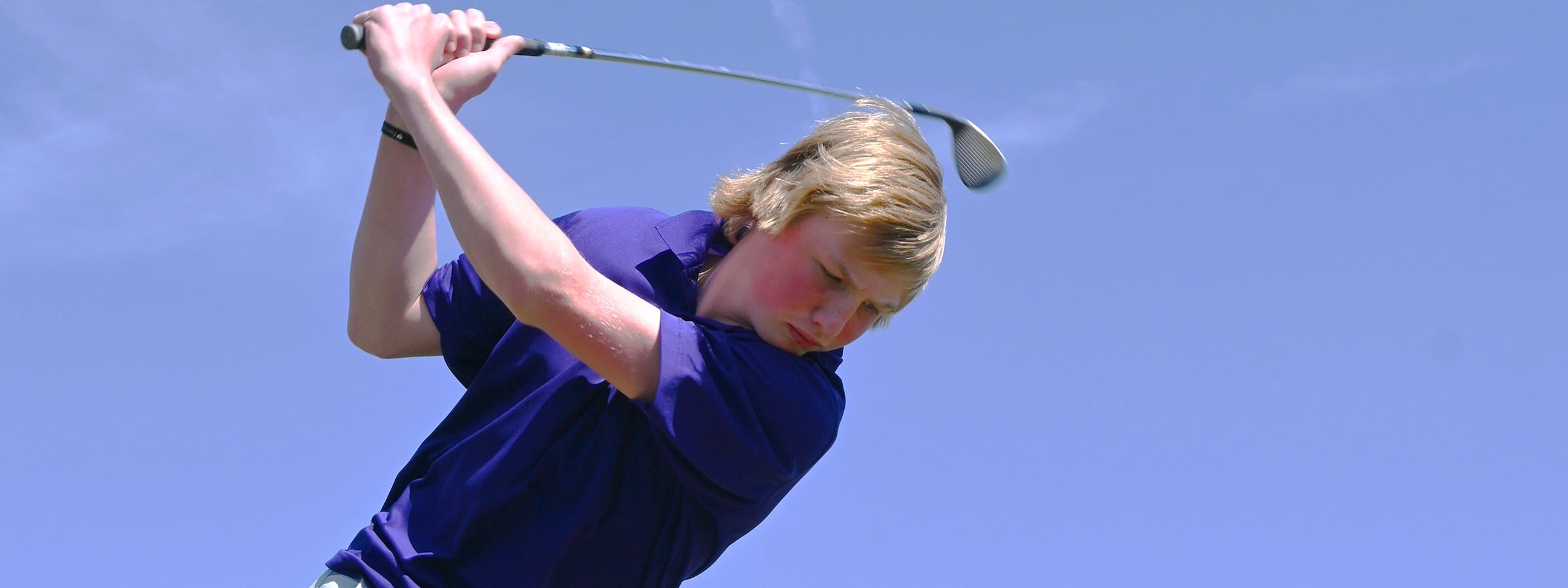 A young man in a golf outfit swings a club, focusing intently on his shot on a sunny golf course.