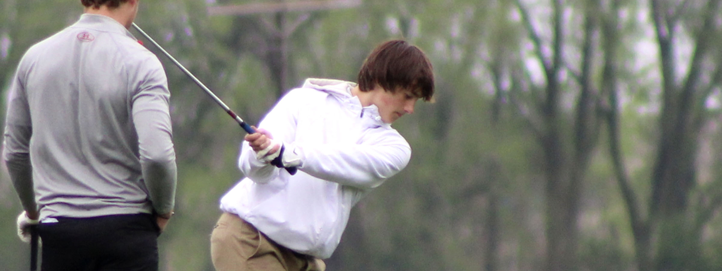 A man swings a golf club as another man watches.