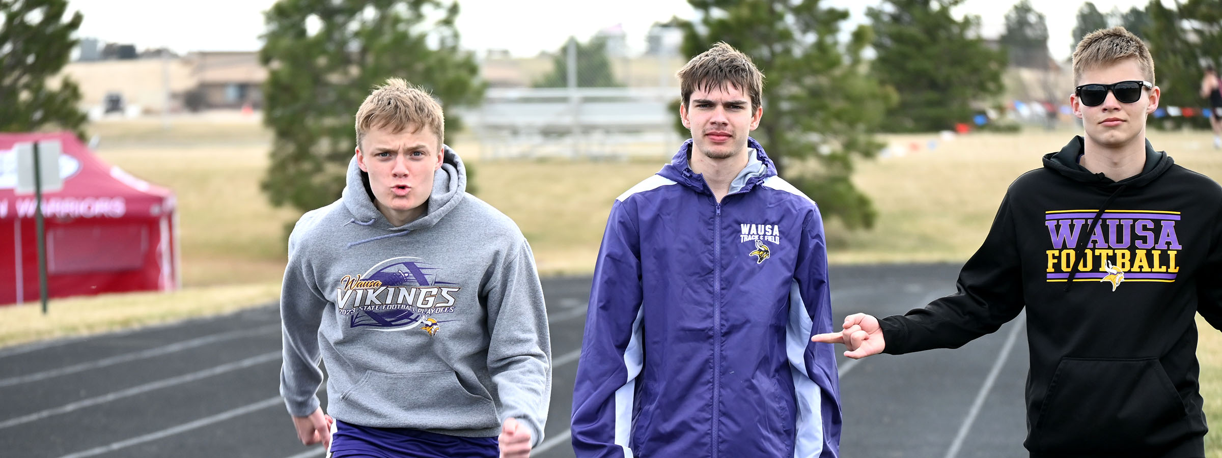 Three young men wearing sweatshirts and sunglasses pose confidently on a track, enjoying a sunny day together.