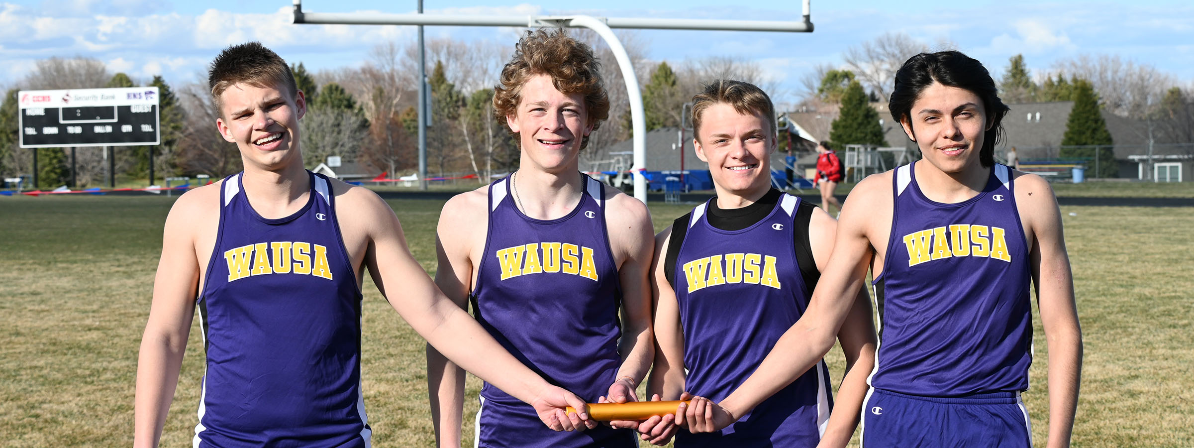 Four boys in purple track suits excitedly hold a baton, ready for a relay race, showcasing teamwork and enthusiasm.