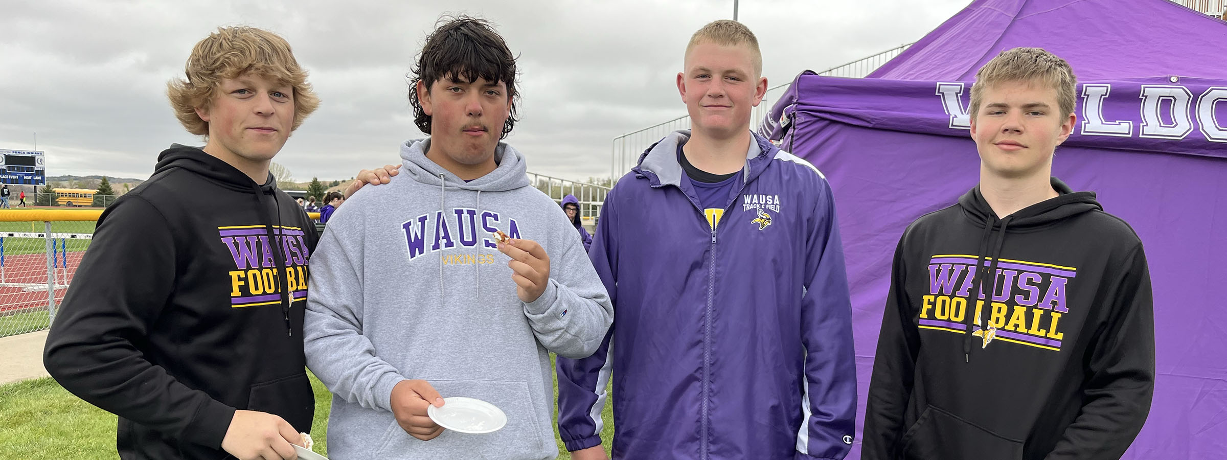 Four young men in purple and white track suits holding a ribbon at a sports event.