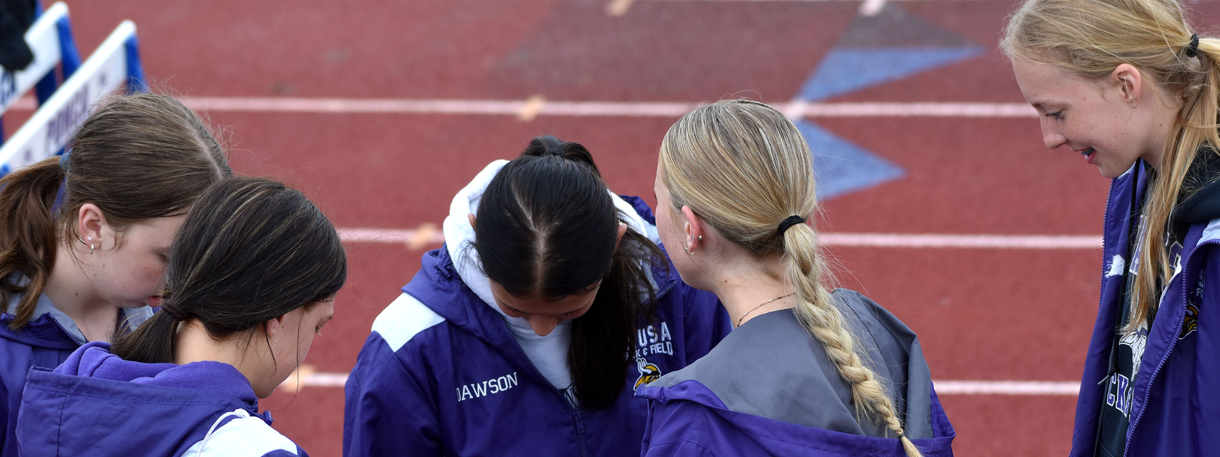 Several girls in purple jackets standing around a track, chatting and laughing together.