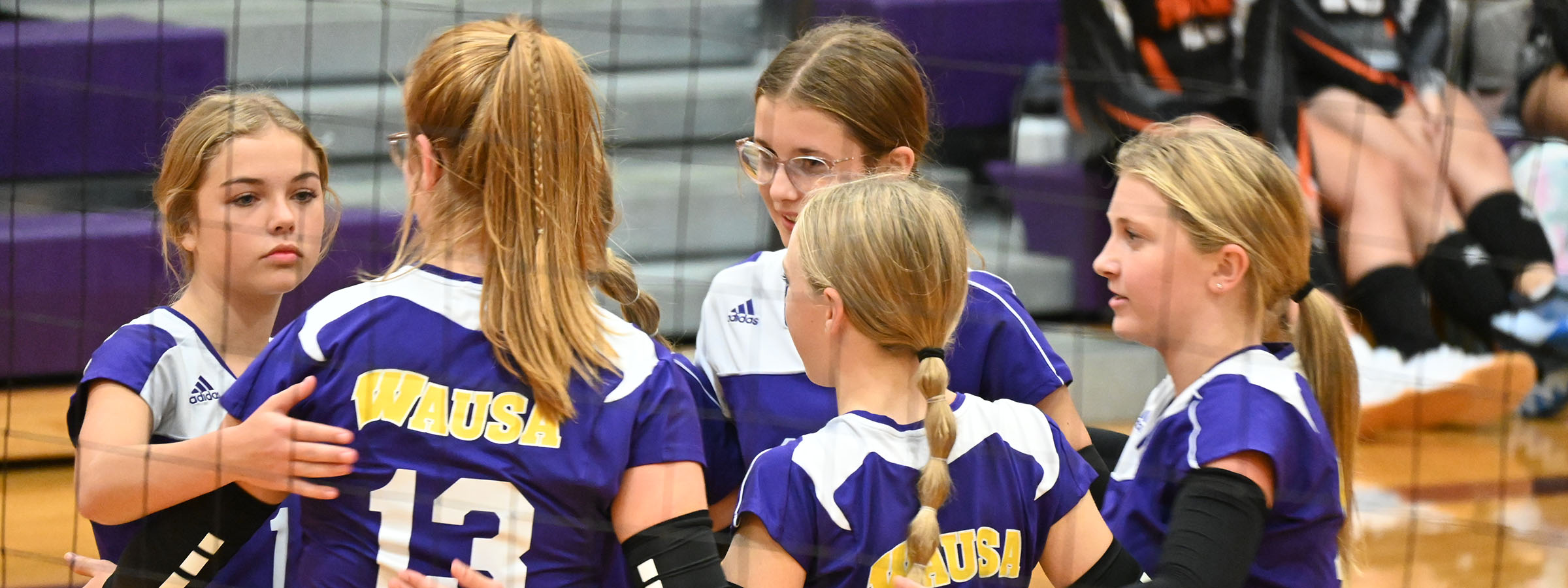 A girls volleyball team huddles together on the court, displaying unity and teamwork before their match.