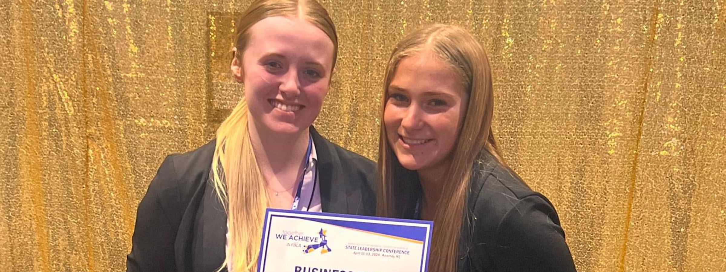 Two women proudly displaying a certificate in front of a gold curtain.