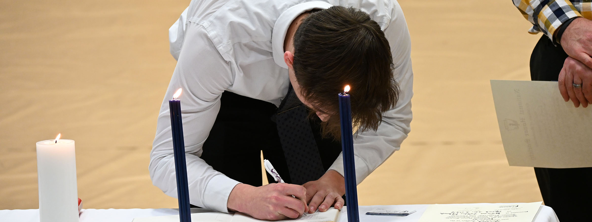 A man focused on signing a document, illuminated by the warm glow of a candle in front of him.