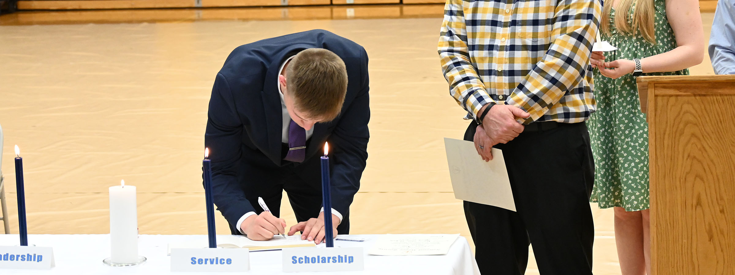 A man focused on signing a document, illuminated by the warm glow of a candle in front of him.