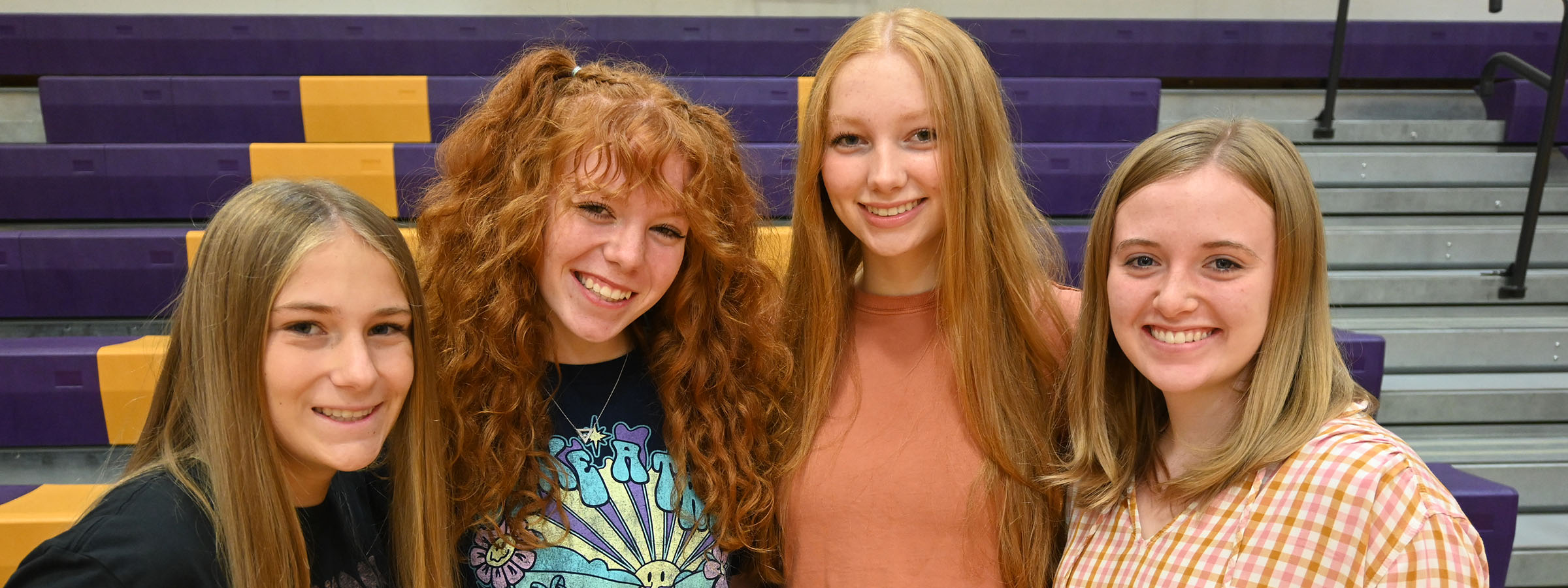 Four girls smiling in front of bleachers at a sports event.