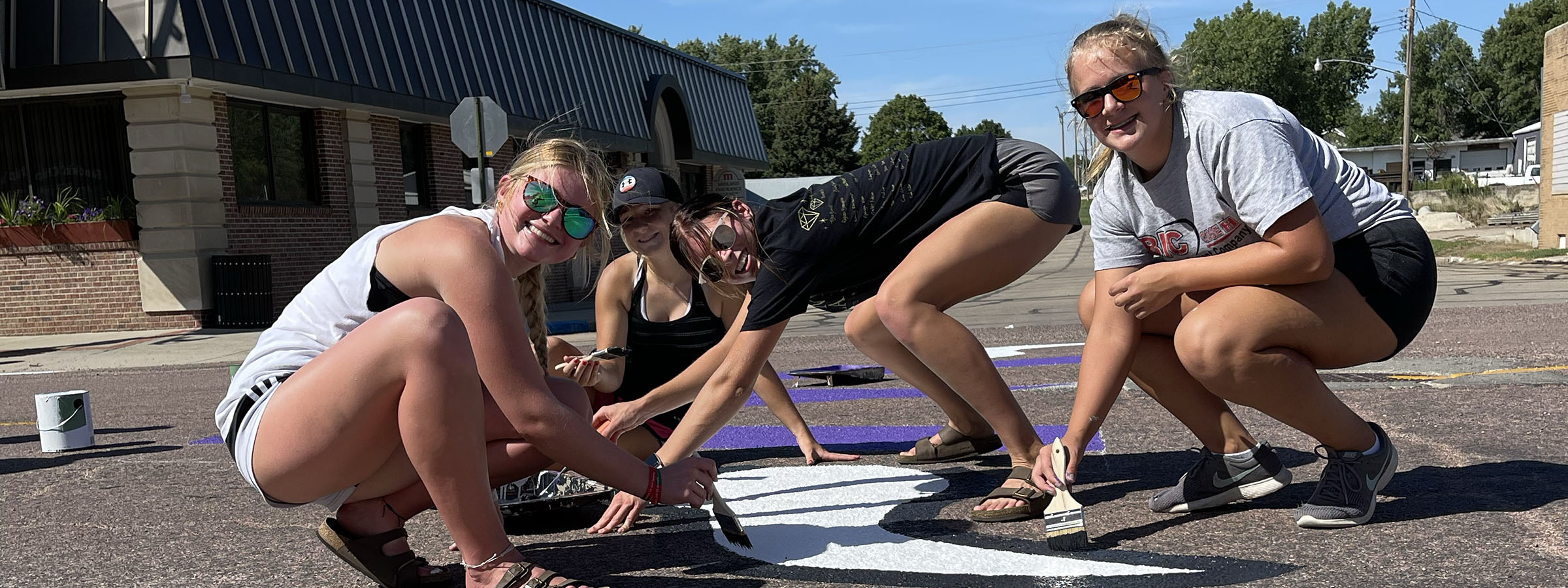 Three women painting a purple and white sign together.